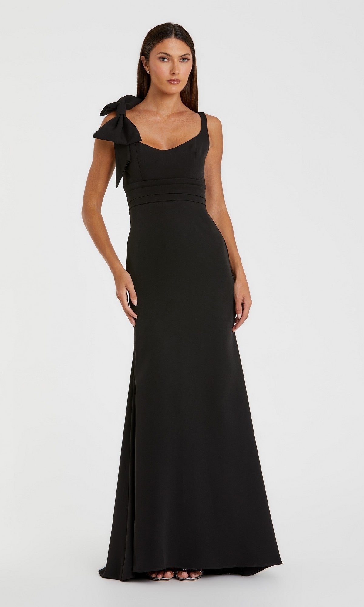 A woman stands against a plain white background wearing the Mac Duggal 49964 long formal dress in black—an elegant, sleeveless gown with a column silhouette and a bow on her left shoulder, epitomizing timeless style.